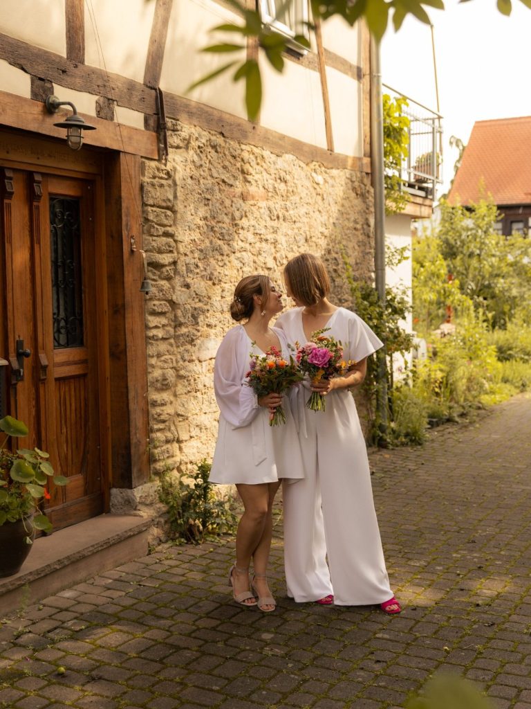 Zwei Bräute stehen auf einem gepflasterten Weg vor einem Fachwerkhaus und halten Blumen.