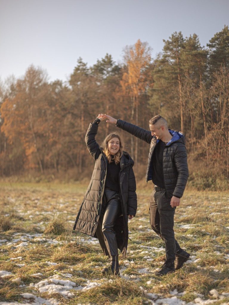 Junges Paar tanzt im Freien, umgeben von herbstlichen Bäumen und schneebedecktem Boden.