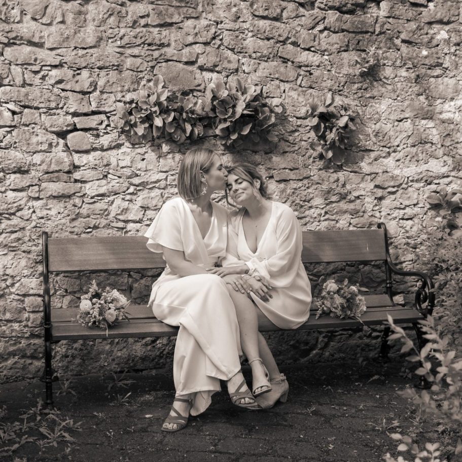 Zwei Frauen küssen sich liebevoll auf einer Bank vor einer Steinmauer.