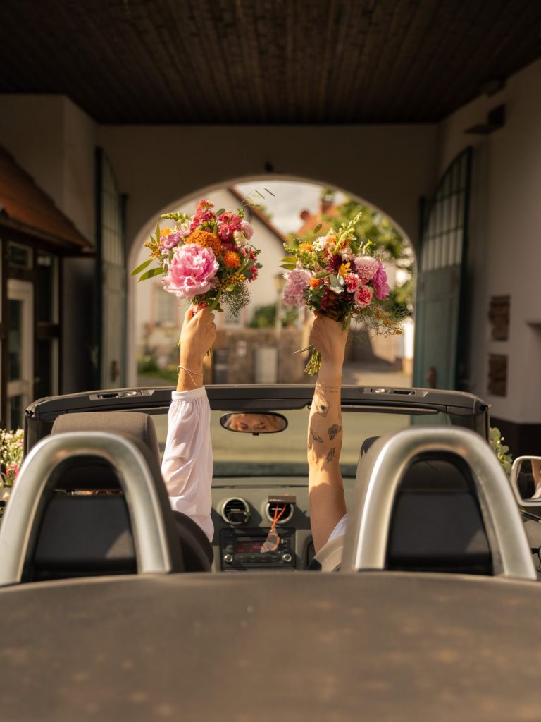 Zwei Bräute halten bunte Blumensträuße aus einem Cabrio heraus.