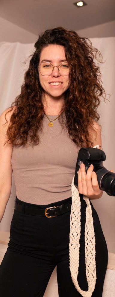 Fotografin mit lockigem Haar hält eine Kamera und lächelt in die Kamera.