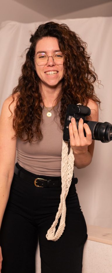 Fotografin mit lockigem Haar, die lachend eine Kamera in der Hand hält.