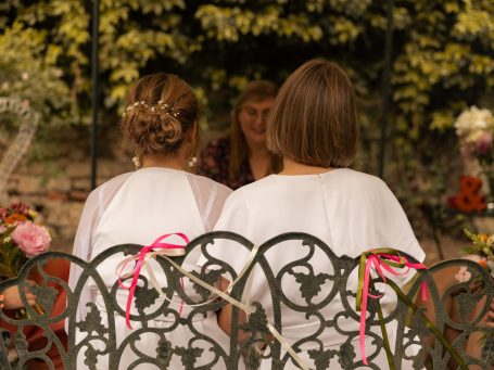 Zwei Frauen in weißen Oberteilen sitzen auf einer Bank und schauen zu einer Person.