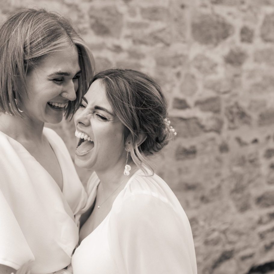 Zwei Frauen lachen herzlich, umarmen sich vor einer steinernen Wand.