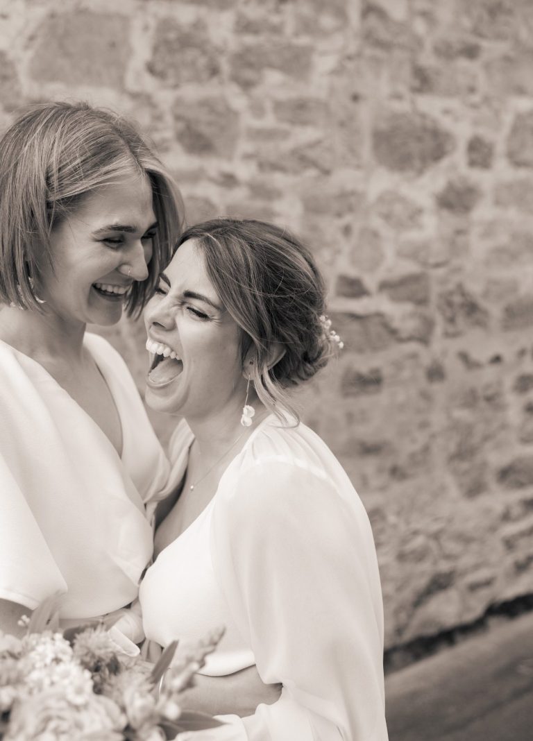 Zwei Frauen in Hochzeitskleidern lachen und umarmen sich vor einer steinernen Wand.