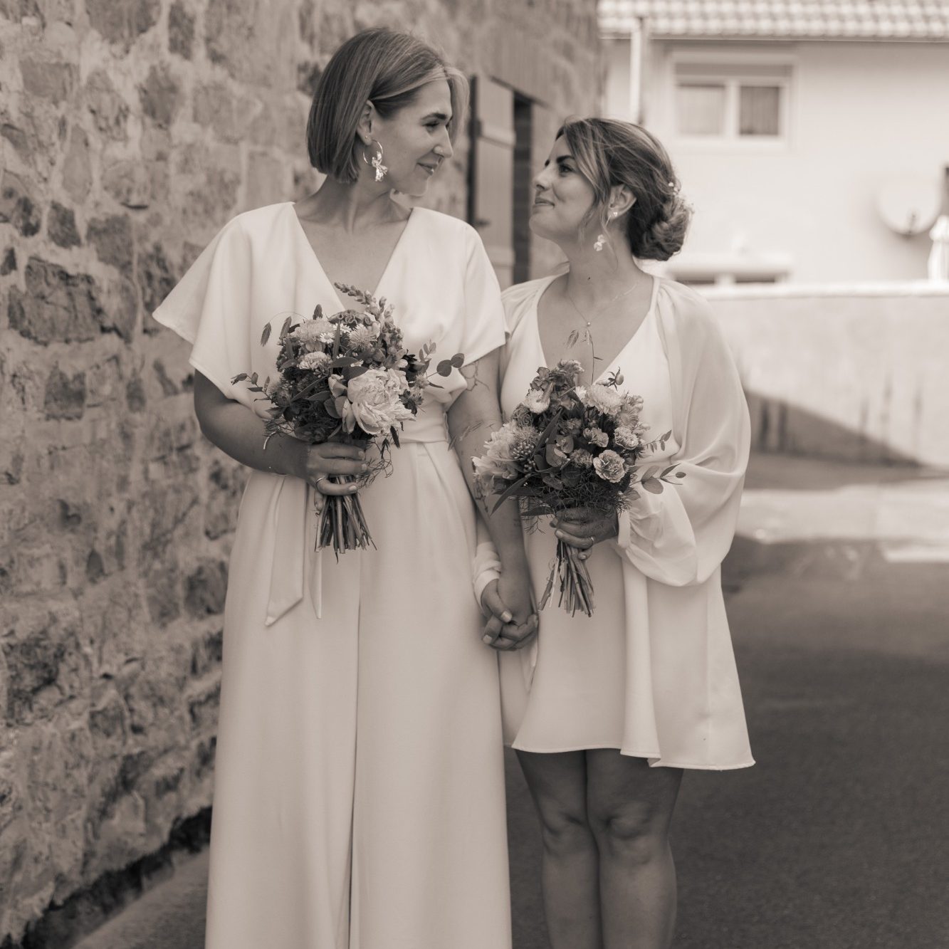 Zwei Bräute in eleganten Kleidern halten Blumensträuße und stehen Hand in Hand.