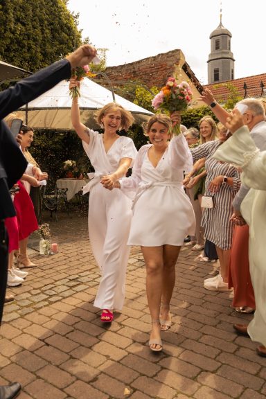 Zwei Frauen in weißen Kleidern laufen fröhlich durch eine Feier, umgeben von Gästen.