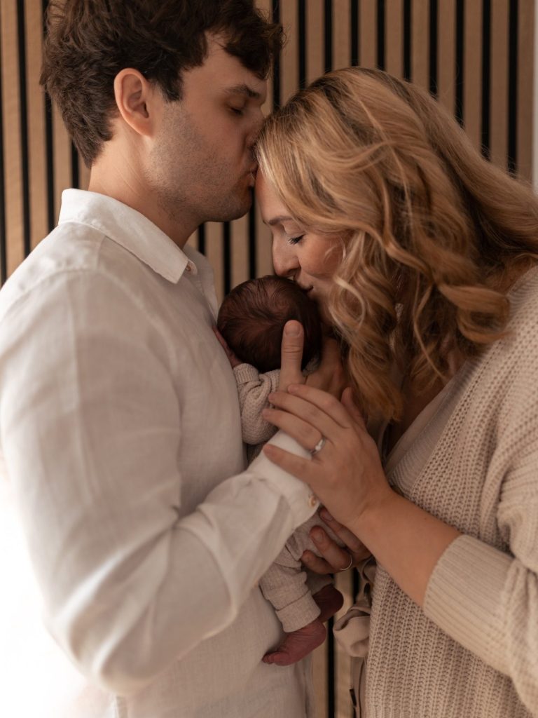 Ein Paar küsst ihr neugeborenes Baby, umgeben von warmem Licht und Holzpaneelen.