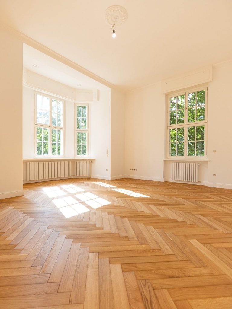 Lichtdurchfluteter Raum mit Parkettboden und großen Fenstern, die Tageslicht hereinlassen.